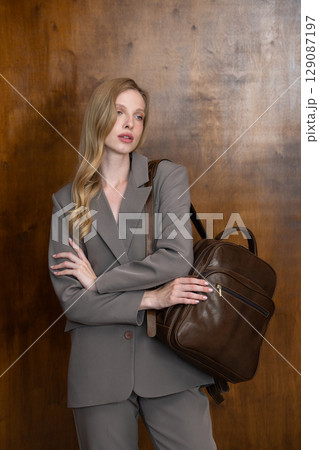 A stylish young woman, long blonde hair, poses in a grey suit, holding a brown leather backpack against a warm wooden background A stylish young woman, long blonde hair, poses in a grey suit, holding a brown leather backpack against a warm wooden background 129087197