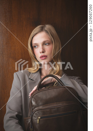 A stylish young woman, long blonde hair, poses in a grey suit, holding a brown leather backpack against a warm wooden background 129087260