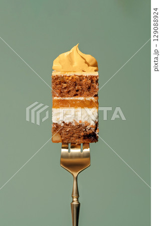 Colorful bento carrot cake slice with orange and mango filling, held by a golden fork, showcasing layers of sponge, cream, and frosting against a soft green background 129088924