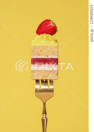 Colorful lemon and strawberry bento cake slice with layers of cream, fruit, and sponge on a golden fork against a vibrant yellow background, showcasing a delightful dessert concept 129088925