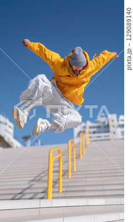 Man in yellow jacket and gray beanie performs dynamic parkour jump against clear blue sky, showcasing agility and energy 129089140