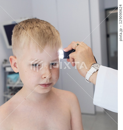 A pediatric dermatologist examining a young boys skin with a dermatoscope in a medical clinic. Skin check up for moles and birthmarks. A pediatric dermatologist examining a young boys skin with a dermatoscope in a medical clinic. Skin check up for moles and birthmarks. 129090624