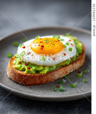 Avocado toast with fried egg on whole grain bread, garnished with chives and chili flakes, served on gray plate Avocado toast with fried egg on whole grain bread, garnished with chives and chili flakes, served on gray plate 129091238
