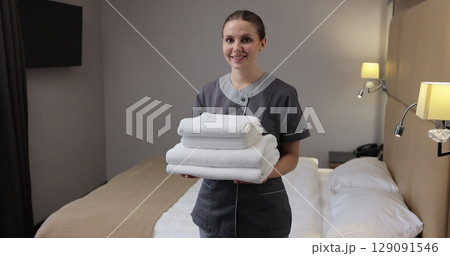 Portrait of young woman housekeeper in uniform holding white towels standing next to bed in modern hotel room smiling to camera. 129091546