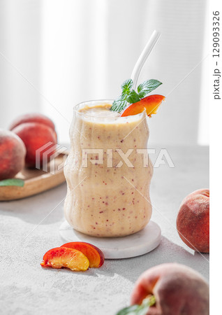Smoothie with peach and banana in a glass on a light background near window with tropical fruits Smoothie with peach and banana in a glass on a light background near window with tropical fruits 129093326