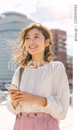 携帯/スマホを手にしながら、携帯/スマホを手にしながら、風に髪をなびかせる笑顔の女性｜光に包まれた都 129093992
