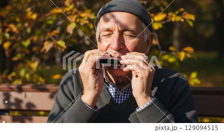 Senior Caucasian man with gray beard wearing knit beanie playing harmonica outdoors in autumn park with golden foliage background 129094515