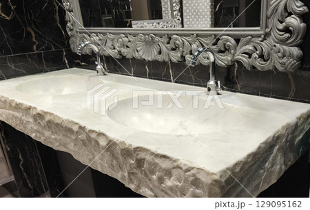 Luxurious marble double sink with raw-edge design and ornate mirror in a black-tiled restroom. Perfect for interior design projects or spa and hotel promotions Luxurious marble double sink with raw-edge design and ornate mirror in a black-tiled restroom. Perfect for interior design projects or spa and hotel promotions 129095162