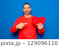 Bearded Hispanic man in his 40s wearing a red sweater holding a red heart as a symbol of romance and health, looking surprised and pointing at himself with his finger. Isolated on a blue background. 129096110