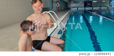 A father helps his son with swimming goggles in a large indoor pool. Concept: strengthening family bonds through training, joint sports, fatherhood. 129098441