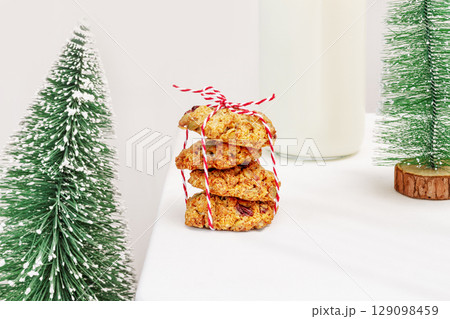 Christmas and New Year still life. Winter holidays food composition with natural handmade cookies with seeds and cranberries, bottle of milk and small new year trees on a white table 129098459