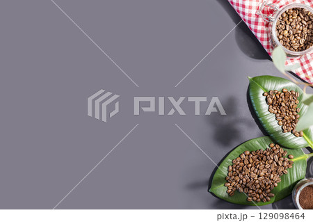 Organic roasted coffee beans on a green leaves and coffee powder in a glass jar on a dark grey background with shadows and copy space. Coffee harvest and making concept. Natural farm coffee 129098464