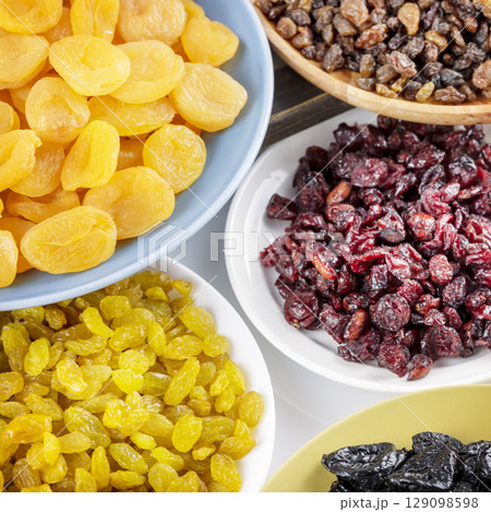 Dry fruits assortment. Dried apricots, cranberries, raisins, and prunes on plates on white background. Healthy snack - mix of organic dry fruits. Vegan and vegetarian products. Closeup Dry fruits assortment. Dried apricots, cranberries, raisins, and prunes on plates on white background. Healthy snack - mix of organic dry fruits. Vegan and vegetarian products. Closeup 129098598