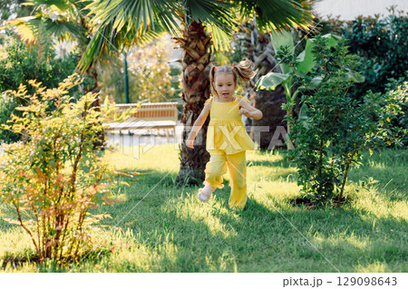 Energetic little girl in yellow dress running joyfully through green garden on a sunny summer day. 129098643
