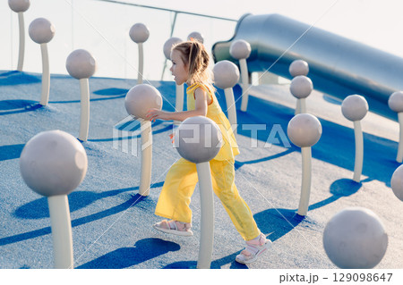 Little girl in yellow exploring modern playground with climbing structures on a sunny summer day. Little girl in yellow exploring modern playground with climbing structures on a sunny summer day. 129098647