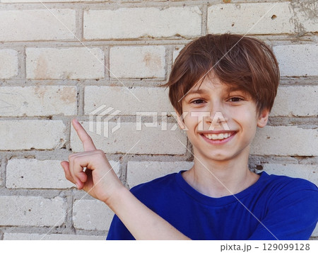 Cute smiling boy in plain blue t shirt looking and pointing hand up in a beige brick wall background 129099128