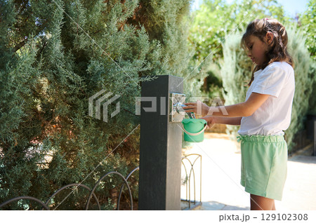 Young Girl Filling a Container From a Garden Water Tap Surrounded by Lush Trees Young Girl Filling a Container From a Garden Water Tap Surrounded by Lush Trees 129102308