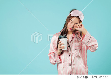 Young woman in pajamas with eye mask holding coffee cup after waking up, she needs more sleep, walking to get some coffee, still needs some sleep, headshots isolated studio blue background Young woman in pajamas with eye mask holding coffee cup after waking up, she needs more sleep, walking to get some coffee, still needs some sleep, headshots isolated studio blue background 129104332