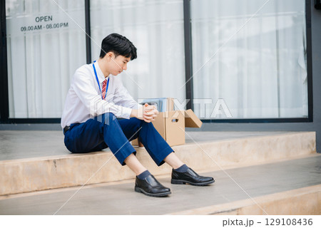 Businessman sitting outside the office with a box of belongings, reflecting on job loss. Capturing the emotional impact and reality Businessman sitting outside the office with a box of belongings, reflecting on job loss. Capturing the emotional impact and reality 129108436