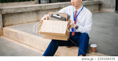 Businessman sitting outside the office with a box of belongings, reflecting on job loss. Capturing the emotional impact and reality 129108439