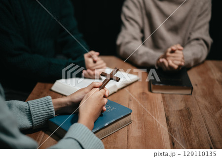 Group of Christians sit together and pray around a wooden table with blurred open Bible pages in their homeroom. Group of Christians sit together and pray around a wooden table with blurred open Bible pages in their homeroom. 129110135