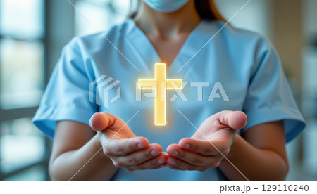 Glowing cross floating above hands of medical worker in blue scrubs and mask, symbolizing faith and hope in healthcare setting 129110240