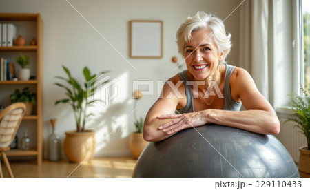 Smiling senior woman with short white hair leaning on exercise ball in bright room with plants and wooden furniture, feeling happy and healthy 129110433