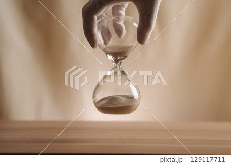 A glove-covered hand holding an hourglass on a wooden surface, symbolizing the flow of time, creativity, and patience in a contemporary minimalist style. A glove-covered hand holding an hourglass on a wooden surface, symbolizing the flow of time, creativity, and patience in a contemporary minimalist style. 129117711