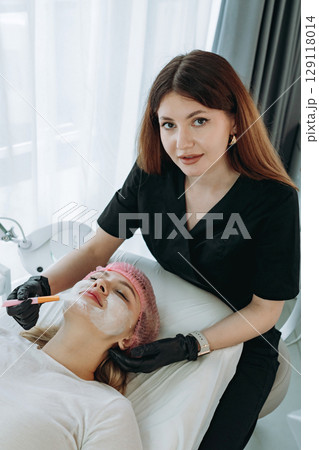 professional aesthetician applies a creamy face mask on young woman face using a brush. Doctor looking at camera professional aesthetician applies a creamy face mask on young woman face using a brush. Doctor looking at camera 129118014