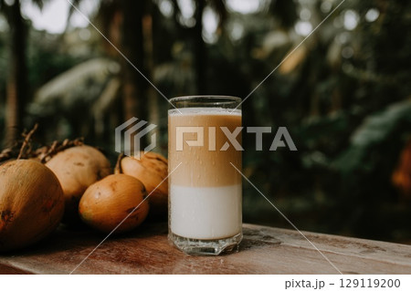 Vietnamese coconut coffee in a tall glass on wooden table, coconuts next to glass. Tropical trees on background Vietnamese coconut coffee in a tall glass on wooden table, coconuts next to glass. Tropical trees on background 129119200