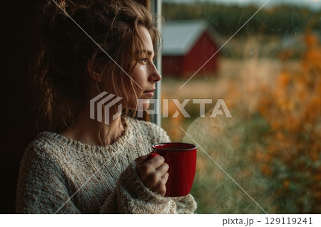 woman wearing knit cozy sweater standing by the window with coffee mug. Autumn rural landscape outside 129119241