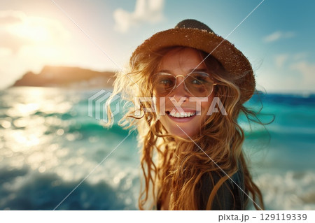 A beautiful young smiling woman on a sunny beach, wearing stylish sunglasses and a wide-brimmed hat. The wind blows through her loose, flowing hair 129119339