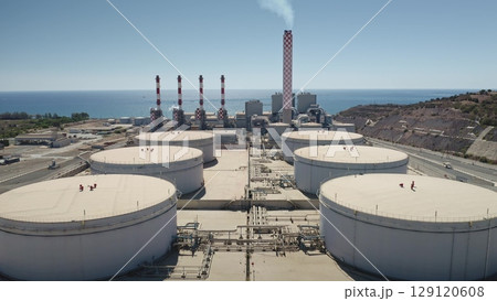 Cyprus: Aerial view of Vasilikos Power Station featuring large white fuel storage tanks and red chimneys emitting smoke along the Cyprus coastline near the Mediterranean Sea. Drone flight Cyprus: Aerial view of Vasilikos Power Station featuring large white fuel storage tanks and red chimneys emitting smoke along the Cyprus coastline near the Mediterranean Sea. Drone flight 129120608