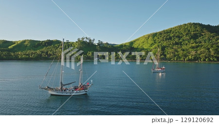 Fiji islands: Two sailboats anchored in calm water near tropical island covered in green lush vegetation, warm glow of the setting sun in Fiji. Wild nature travel landscape. Aerial drone flight Fiji islands: Two sailboats anchored in calm water near tropical island covered in green lush vegetation, warm glow of the setting sun in Fiji. Wild nature travel landscape. Aerial drone flight 129120692