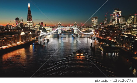United Kingdom, London: Breathtaking aerial view of London at night, illuminated Tower Bridge, The Shard, and cityscape reflecting on the River Thames with boat, creating mesmerizing urban nightscape United Kingdom, London: Breathtaking aerial view of London at night, illuminated Tower Bridge, The Shard, and cityscape reflecting on the River Thames with boat, creating mesmerizing urban nightscape 129120778