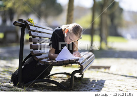 Young student writing on notebook lying on bench in park 129121198