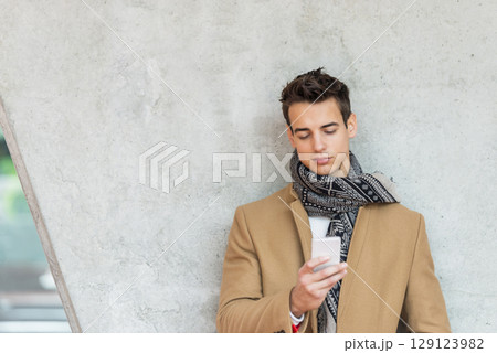 Stylish man texting on smartphone against concrete wall 129123982