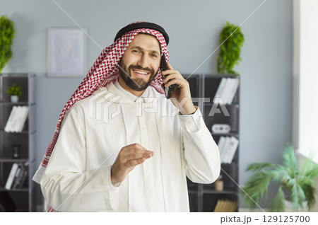 Arab business man in traditional dishdasha dress standing in office talking by mobile phone. Arab business man in traditional dishdasha dress standing in office talking by mobile phone. 129125700