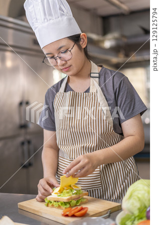 A young Asian woman placing slice of cheese on homemade sandwich. A young Asian woman placing slice of cheese on homemade sandwich. 129125794