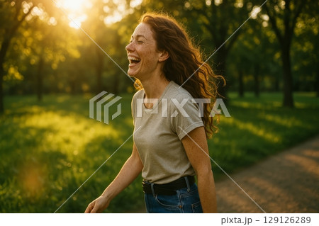 Cheerful woman with freckles and red curly hair laughing while walking in green park during sunset on warm summer evening 129126289