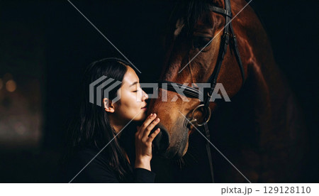 A young woman gently kisses a horse's face at sunset, capturing an intimate moment of trust and connection. The soft backlight highlights their bond in a serene countryside setting 129128110