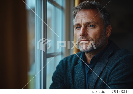 Close-up of thoughtful mature man looking out the window. Natural side light. Thoughtful expression. Inner reflection. Cozy interior. Soft tones. Lifestyle, emotions, copy space. Plenty of text space 129128839