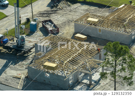 Aerial view of lifting crane and builders working on unfinished residential house with wooden roof frame structure under construction in Florida suburban area 129132350