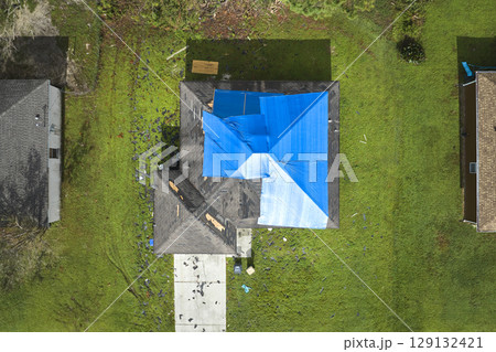 Aerial view of damaged in hurricane Ian house roof covered with blue protective tarp against rain water leaking until replacement of asphalt shingles 129132421