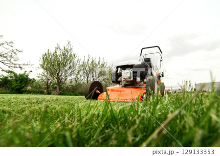 Gardening work tools. Wide angle view of orange electric lawn mower, wheels, motor on bright lush green grass at backyard. Rotary lawn mower machine cut lawn. Professional lawn care service. Gardening work tools. Wide angle view of orange electric lawn mower, wheels, motor on bright lush green grass at backyard. Rotary lawn mower machine cut lawn. Professional lawn care service. 129133353