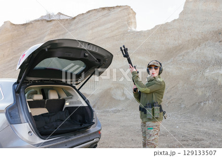 Female Soldier Holding Shotgun near Car with Open Trunk in Desert Area 129133507