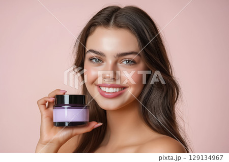 Beauty portrait of a young woman with a neat hairstyle with glowing skin holding a jar of cream on a neutral beige background 129134967
