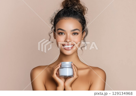 Beauty portrait of a young woman with a neat hairstyle with glowing skin holding a jar of cream on a neutral beige background 129134968