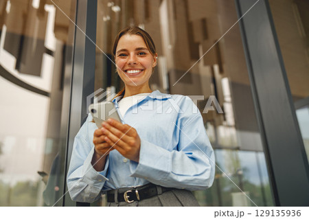 A Young Professional is happily Smiling while Using a Smartphone Outside a Modern Building 129135936