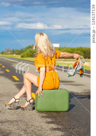 Blonde woman is sitting on a retro suitcase on a rural highway. Blonde woman is sitting on a retro suitcase on a rural highway. 129137139